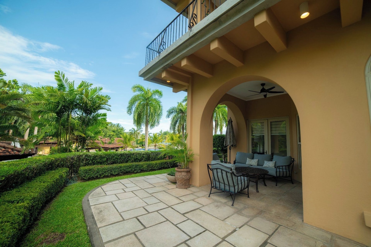A covered outdoor patio with a dining set, a ceiling fan, and a view of a pool and palm trees.