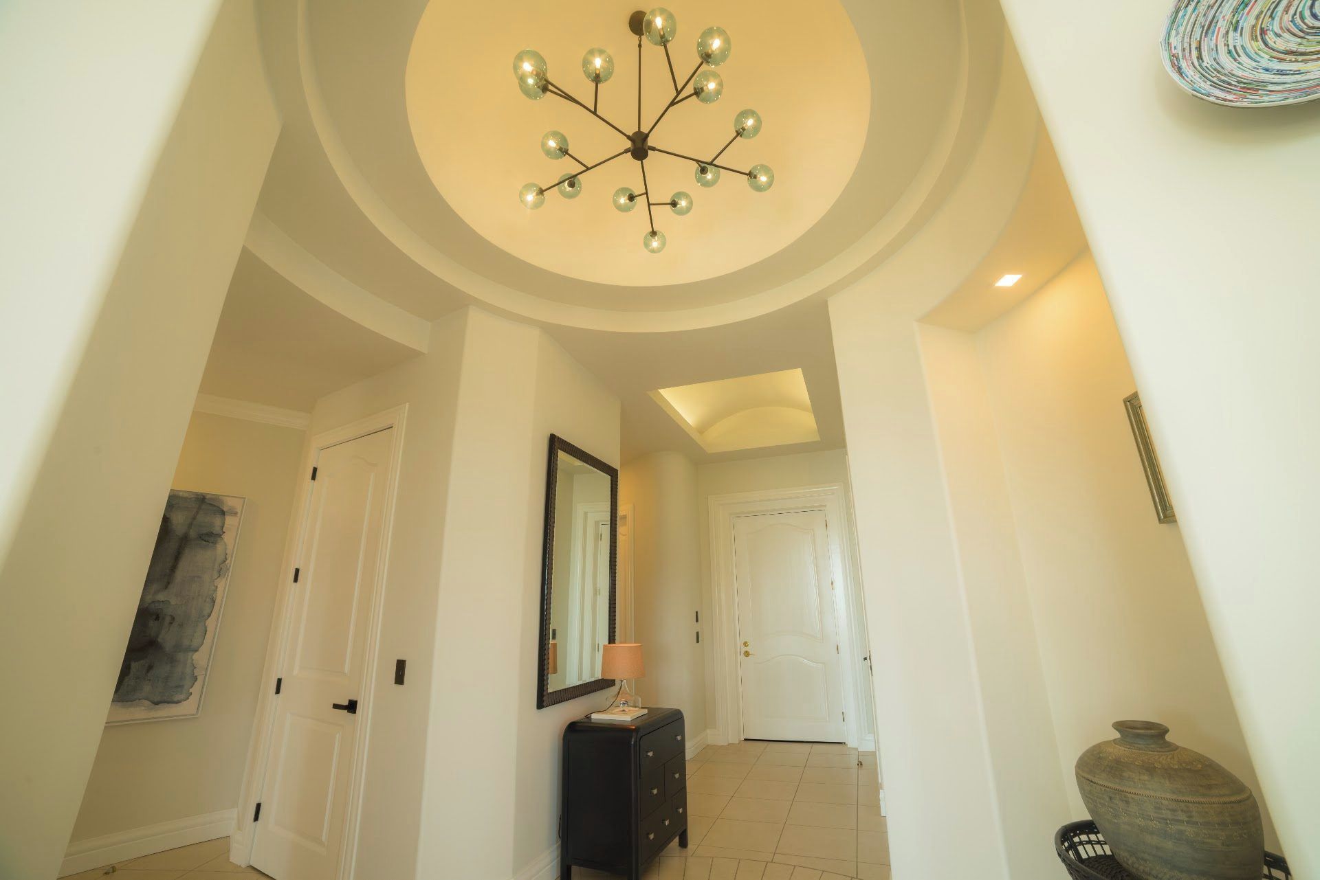 A hallway with a modern chandelier, a mirror, and a vase on a stand.