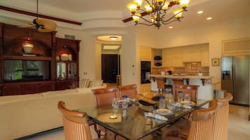 A dining area with a glass table, striped chairs, a chandelier, and a kitchen in the background.