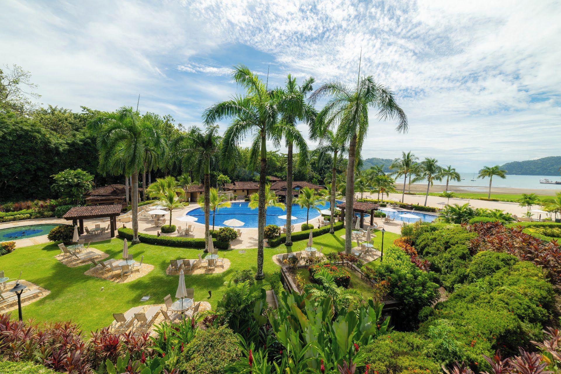 Un resort con piscina central, palmeras y vista al mar.