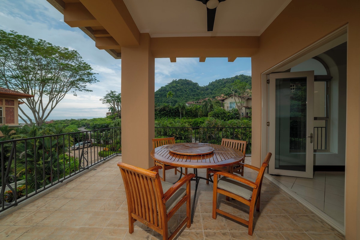 A covered outdoor area with a round wooden table and chairs, a glass door, and a view of a lush green landscape.