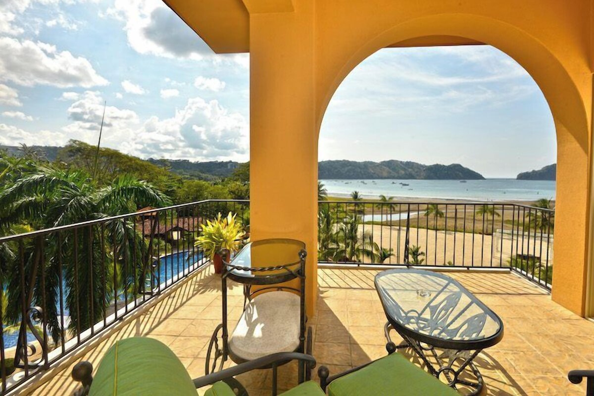 A balcony with a view of the beach, a pool, and a dining area with chairs and a table.