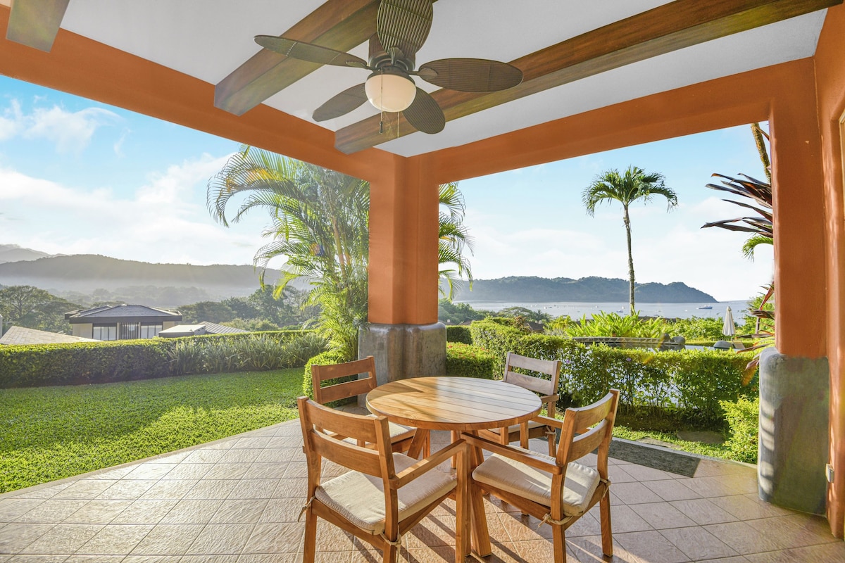 A covered patio with a table and chairs, overlooking a landscaped garden and a view of the sea.