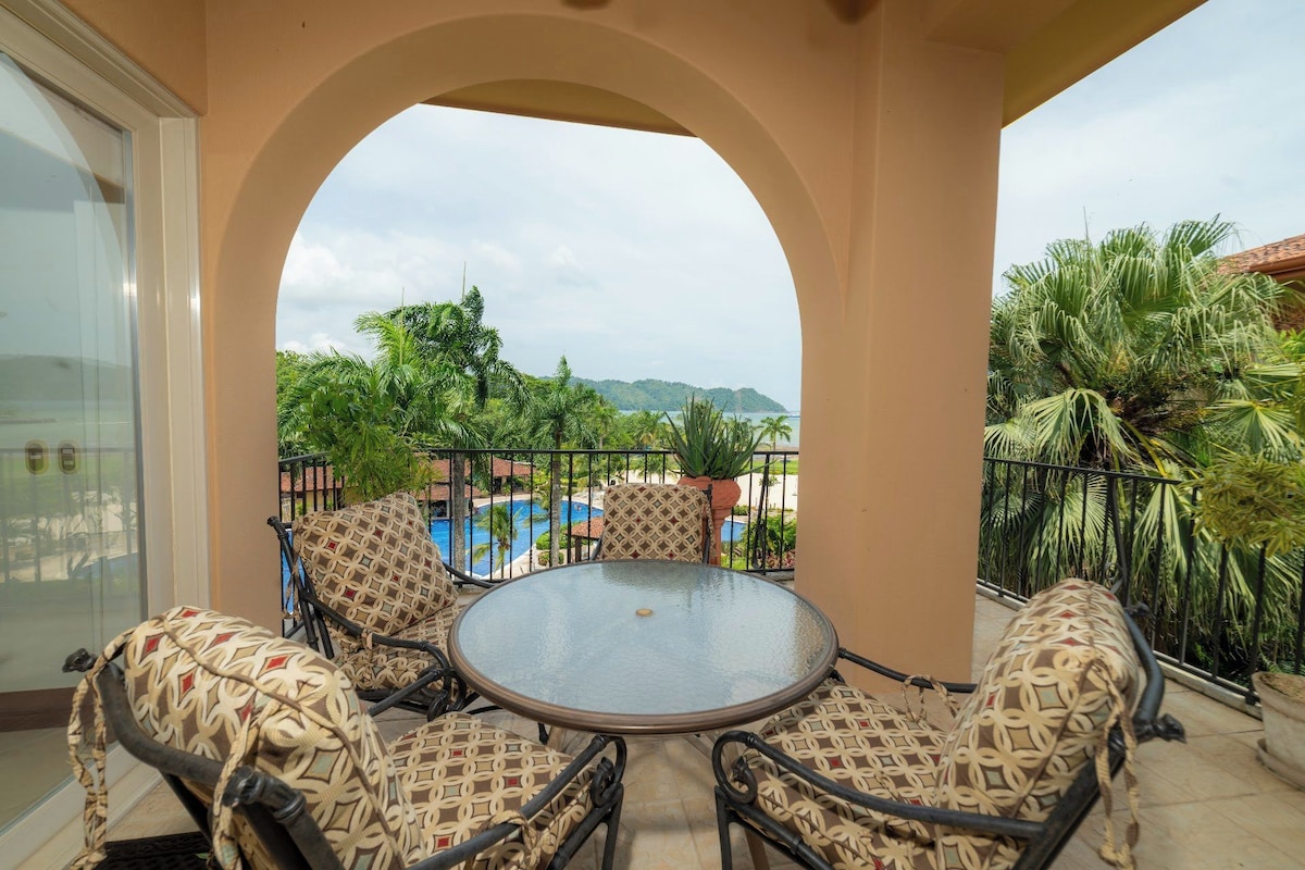 A covered outdoor seating area with a glass table and patterned chairs, overlooking a pool and lush greenery.
