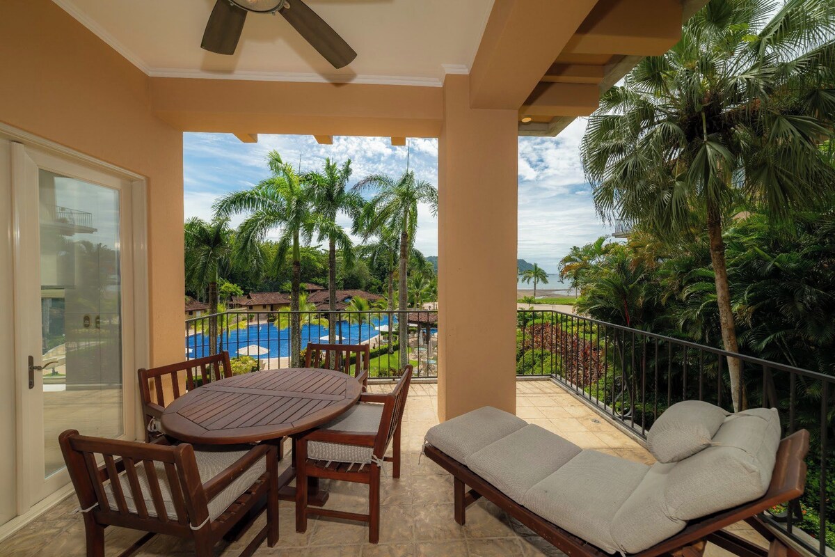 A balcony with a table and chairs, a lounge chair, and a view of a swimming pool and palm trees.