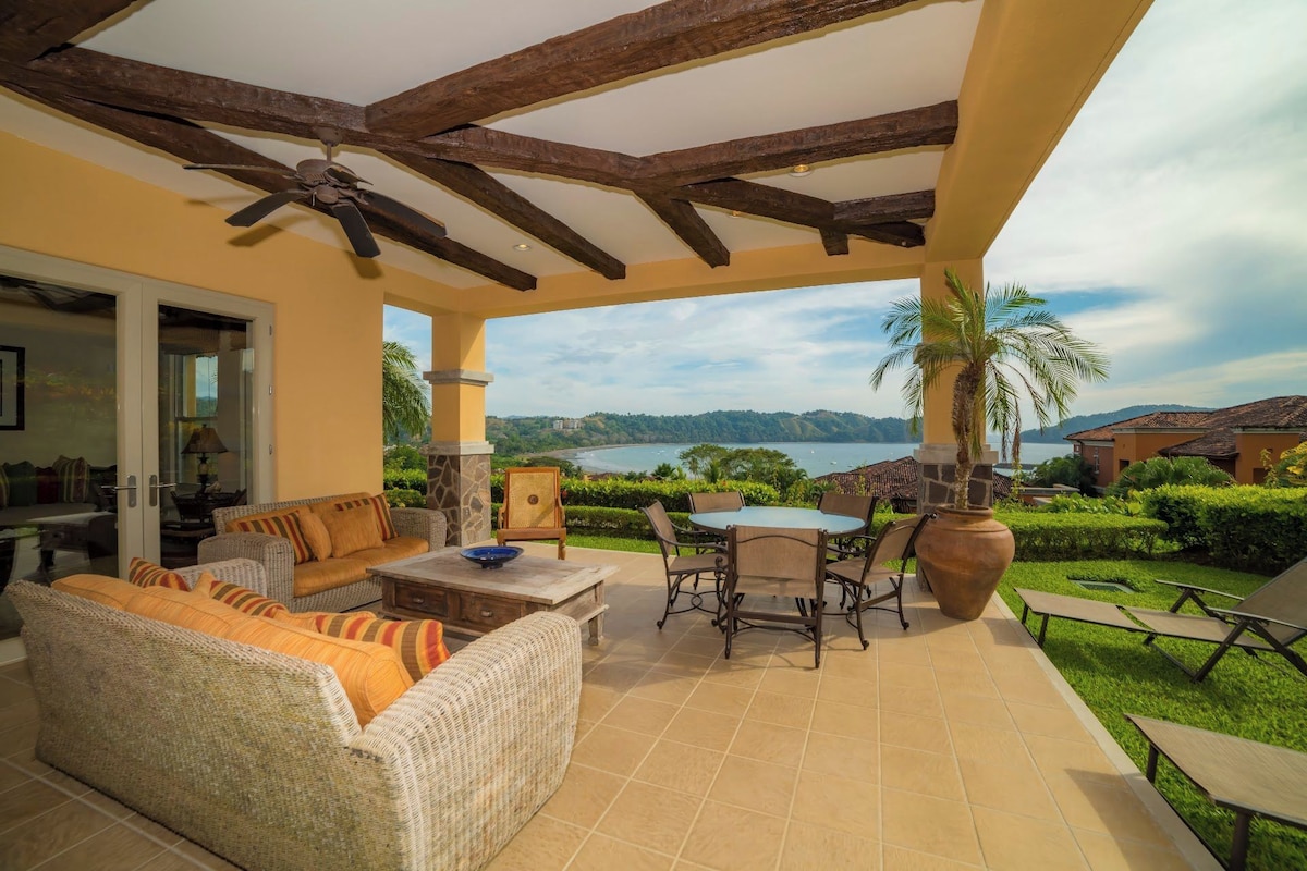 A covered outdoor seating area with a table and chairs, a sofa, and a view of a lake and hills.