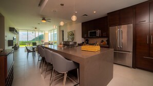 A modern kitchen with a large island, bar stools, stainless steel appliances, and a view of the outdoors. - Montecielo 3E 3bdrs Ocean View in Los Suenos (Jacó)