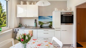 Una cocina moderna con armarios blancos, un lavamanos, una estufa y un refrigerador. Se ve un ventanal con vistas a la vegetación.