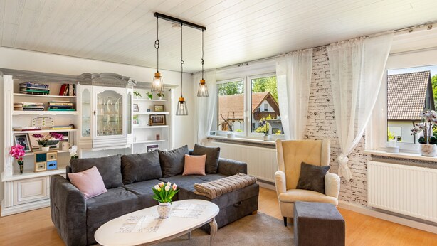 Una sala de estar con sofá, sillón y una mesa de centro, una estantería y una ventana con vista a una casa.