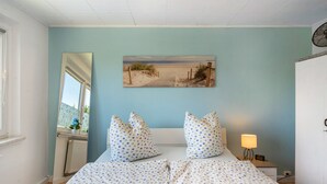 Un dormitorio con cama, una mesita de noche con lámpara, cómoda, ventana con vista y un cuadro de una escena de playa en la pared.