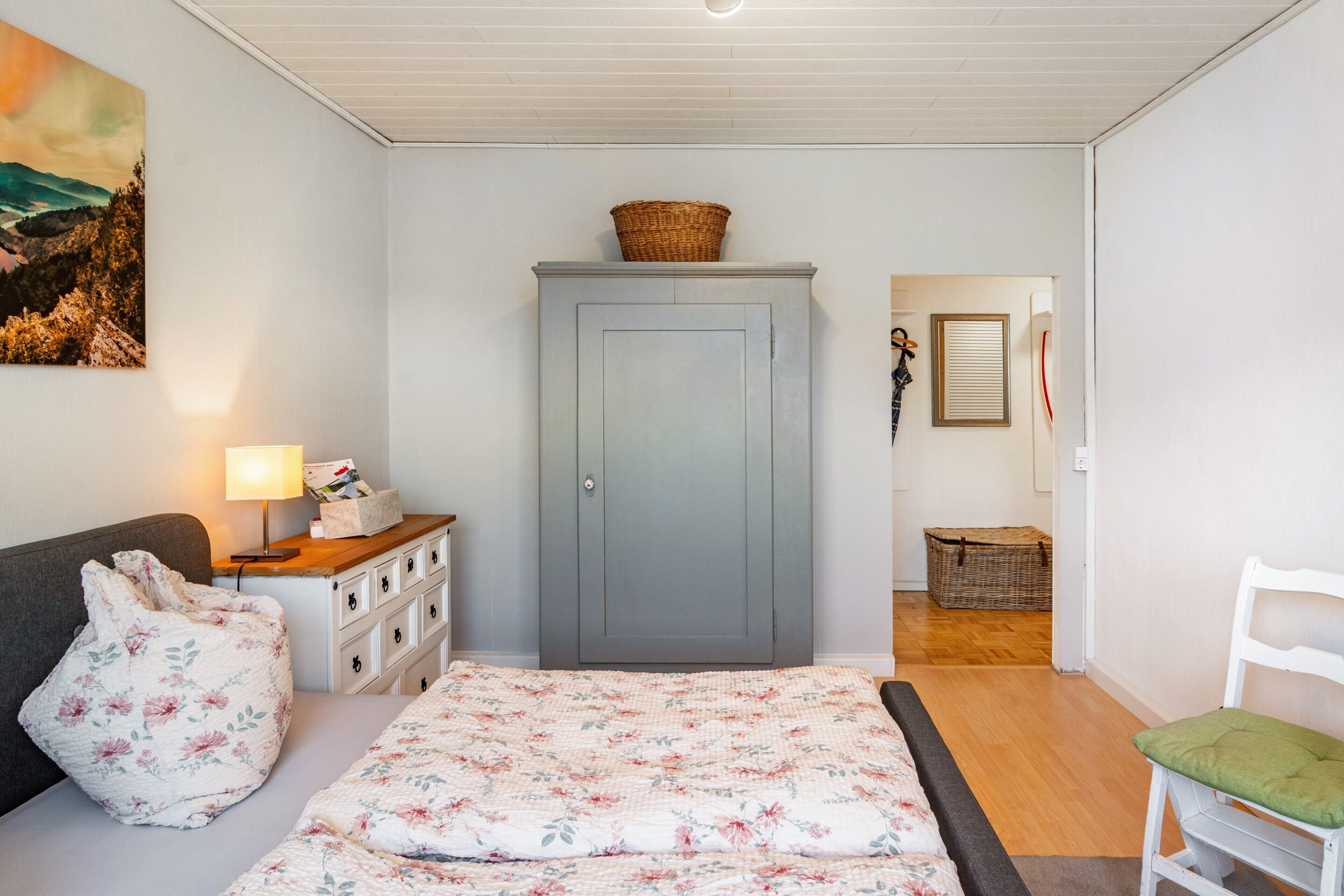 Un dormitorio con cama, cómoda, silla, lámpara, armario y un cuadro en la pared.