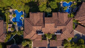 An aerial view of a residential area with multiple swimming pools, a central building with a distinctive roof, and surrounding greenery. - Veranda 3G 2 bdr Garden View in Los Suenos (Jacó)