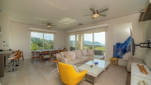 A spacious living area with a yellow armchair, a beige sofa, and a coffee table. There are ceiling fans, large windows offering a view of the outdoors, and a dining area with wooden furniture. - Miramar 5D 3 bdr Ocean View in Los Suenos (Jacó)