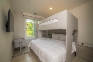 Una cama litera con edredón blanco, una mesita de noche, un ventanal pequeño y un televisor montado en la pared.