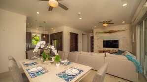 A modern dining area with a wooden table set for four, a ceiling fan, recessed lighting, and a living area with a sofa and a flat-screen TV. - Vista Los Suenos by Stay in Costa Rica (Jacó)