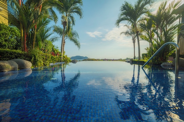 An infinity pool with clear blue water, surrounded by lush greenery and palm trees, with a view of distant hills.