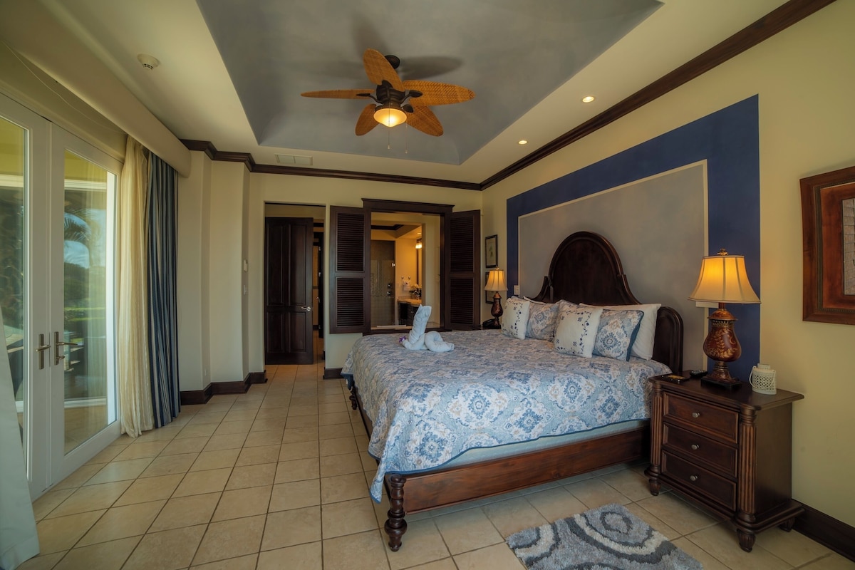 A bedroom with a large bed, a ceiling fan, a nightstand, and a view of the bathroom.