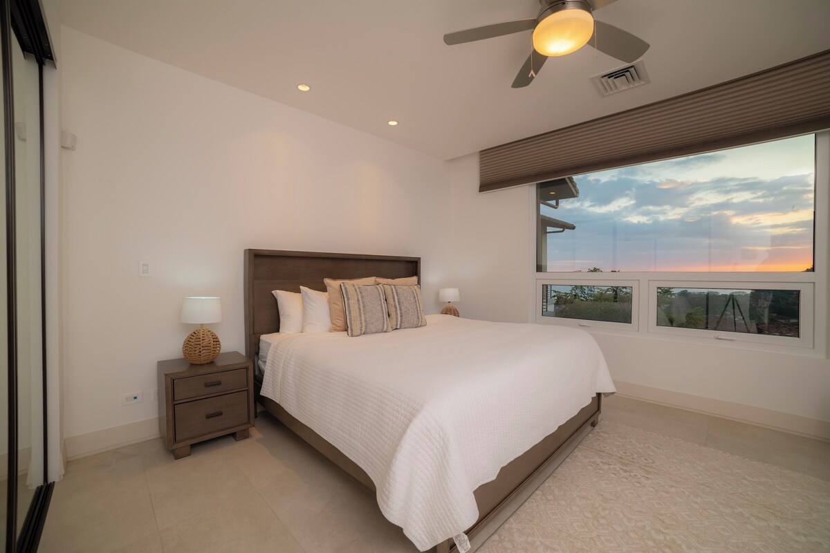 A bedroom with a large bed, a nightstand, a lamp, a ceiling fan, and a window with a view of the sky.