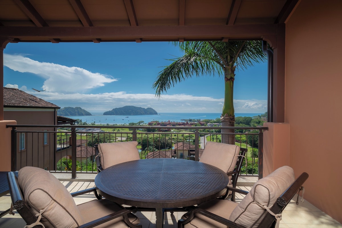 A balcony with a round table and four chairs, overlooking a coastal landscape with a palm tree and a view of the sea.