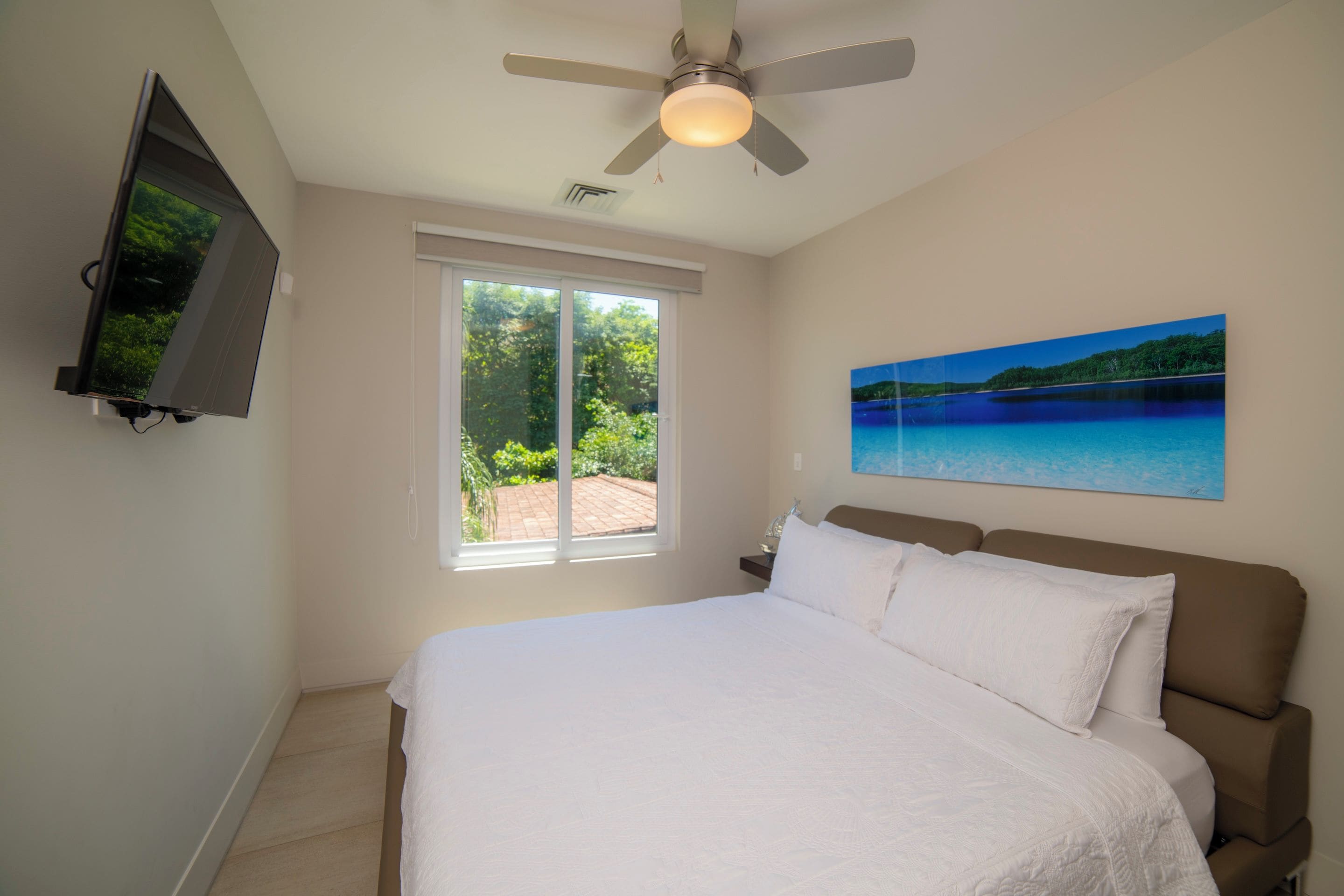 Quarto com cama, ventilador de teto, televisão na parede, janela com vista para o verde e uma pintura de paisagem.