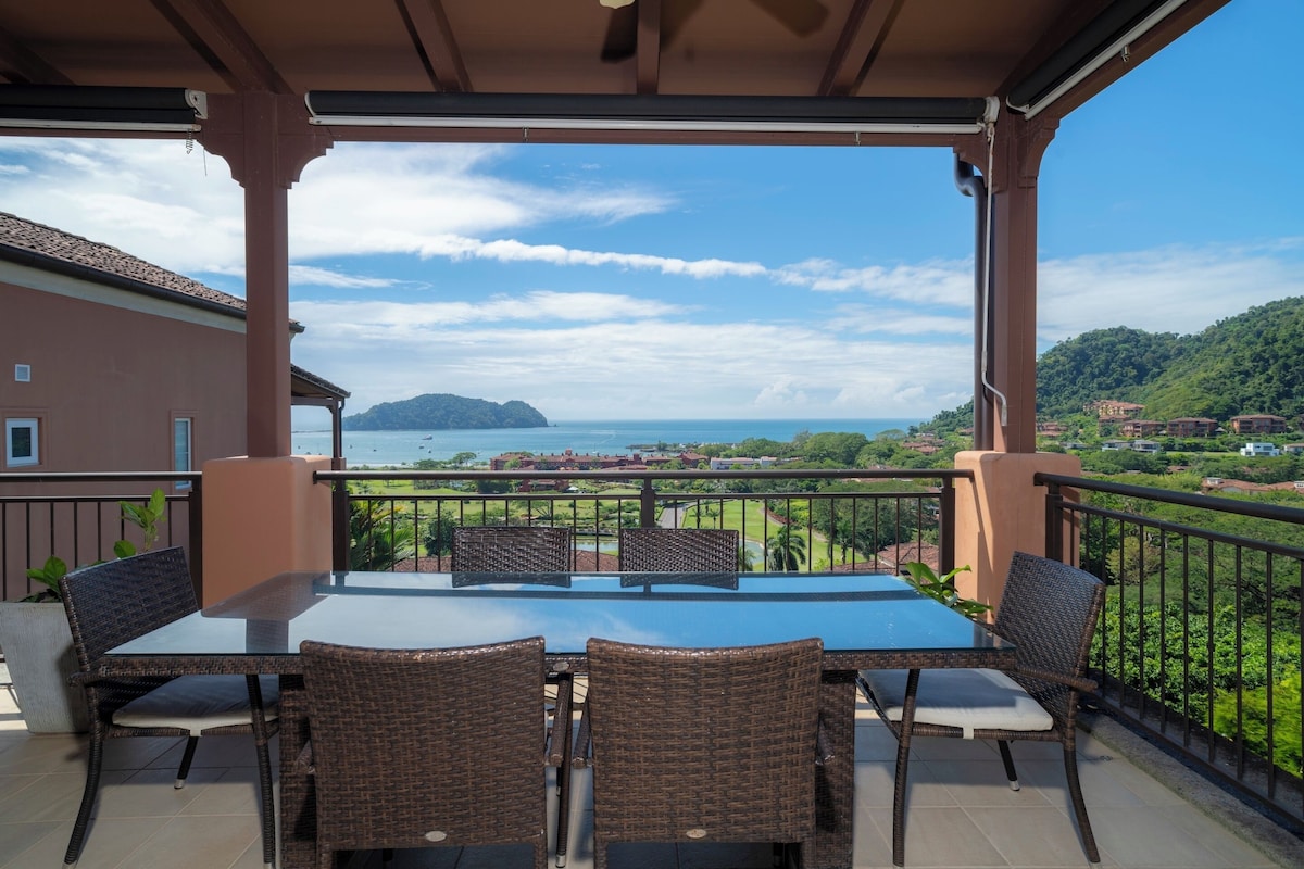 A covered outdoor seating area with a table and chairs, overlooking a coastal landscape with a view of the sea and distant buildings.
