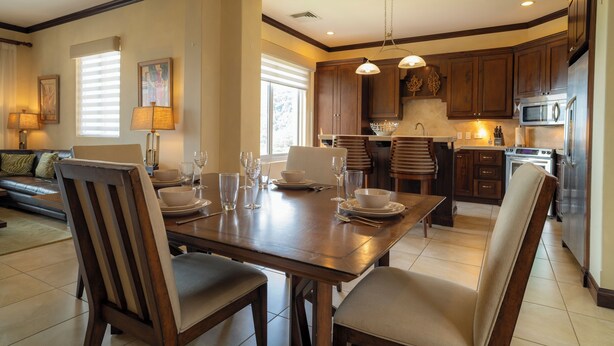 A dining area with a wooden table set for four, surrounded by chairs, in a room with a kitchen in the background. - Vista Los Suenos by Stay in Costa Rica (Jacó)