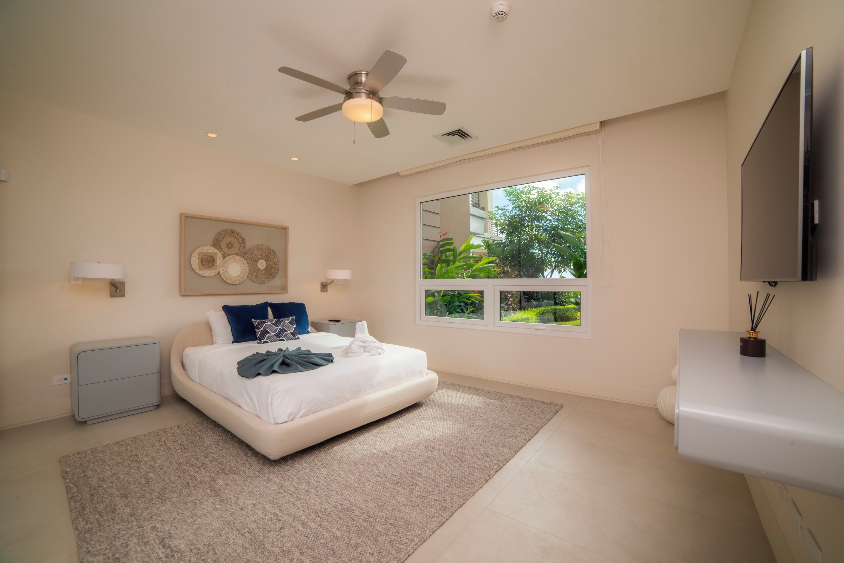 Quarto moderno com cama, ventilador de teto, janela com vista e TV na parede.