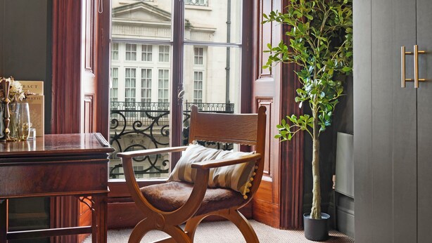 A room with a wooden chair, a desk, a potted plant, and a view of a building through a window with curtains. - Hertford Flat 7 (London)
