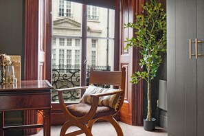 A room with a wooden chair, a desk, a potted plant, and a view of a building through a window with curtains. - Hertford Flat 7 (London)