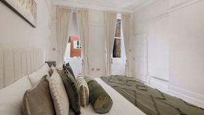 A bedroom with a large bed, beige curtains, a chandelier, and a view of a brick building. - Hertford Flat 8 (London)