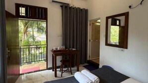 Una habitación con cama, escritorio y silla. Cuenta con ventana con cortinas, una puerta y vista a follaje exterior.