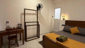 Habitación de hotel con cama, escritorio, silla, ventilador de pie y una mesita con hervidor y tazas.