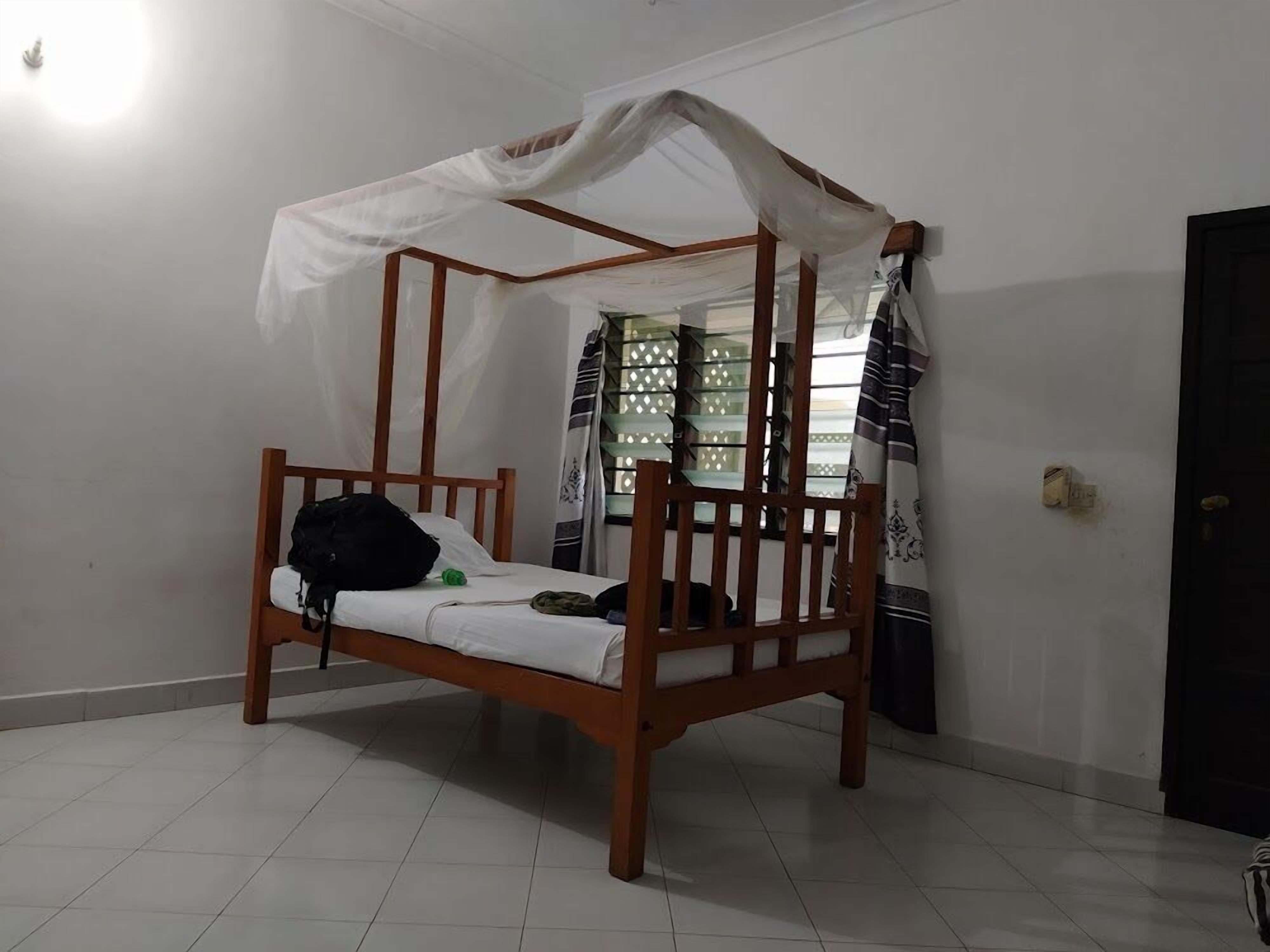 A single wooden bed with a canopy, a small black bag on the bed, and a window with patterned curtains.