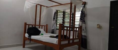 A single wooden bed with a canopy, a small black bag on the bed, and a window with patterned curtains.