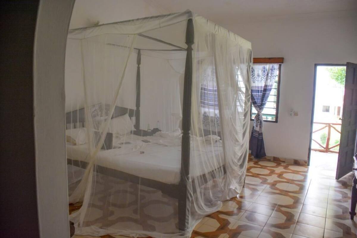 A room with a bed under a mosquito net, a tiled floor, and a window with curtains.