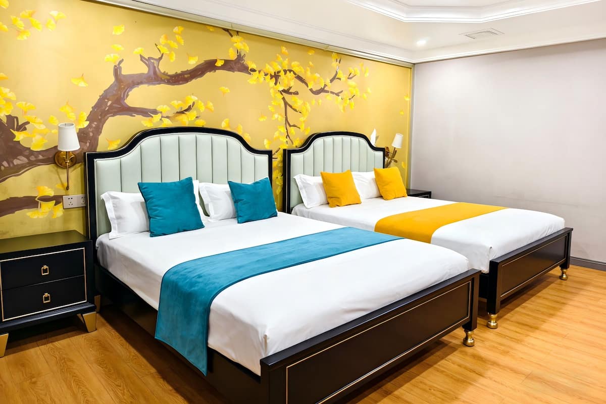 Ein Hotelzimmer mit zwei Betten, jeweils mit weißer Bettwäsche und blauen und gelben Akzenten. Das Zimmer verfügt über eine gelbe Wand mit einem Baum-Wandbild und einen Holzboden.