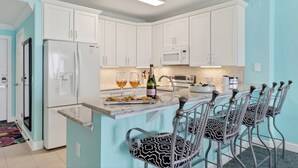 Une cuisine avec des meubles blancs, un réfrigérateur, un four à micro-ondes et un coin bar équipé de chaises et d’une bouteille de champagne.