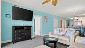 Un salon avec un mur de couleur turquoise, un meuble de télévision noir, un canapé beige et un ventilateur de plafond.