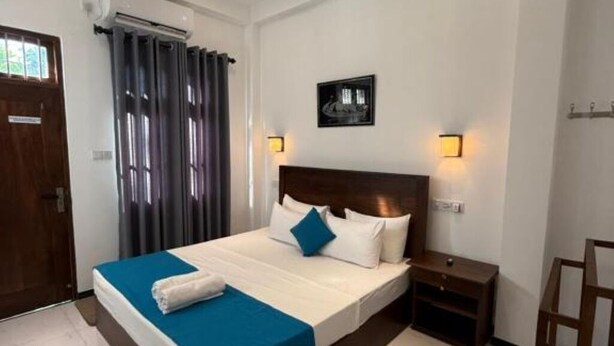 A hotel room with a large bed, a wooden nightstand, a wall-mounted lamp, and a door with a window. - Devon Surf  Inn : Deluxe Double Room (Dikwella)