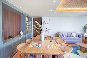 A modern living room with a wooden dining table, a grey sofa, and a bookshelf.