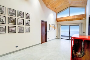 A spacious hallway with framed black and white photographs on the walls, a wooden ceiling, and large windows offering a view of the sky.
