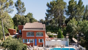 Ein zweistöckiges Gebäude mit Ziegeldach, umgeben von Bäumen und einem Swimmingpool mit Liegestühlen.