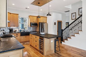 A modern kitchen with wooden cabinets, a black countertop, stainless steel appliances, and a black staircase with a railing. - Modern Home + Firepit + Balcony + Near East Austin | Chestnut Ember by AvantStay (Austin)