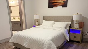 Un dormitorio con una cama grande, dos mesitas de noche con lámparas, una mesita de noche adicional con lámpara, un cuadro colgado en la pared y una puerta que da al baño.