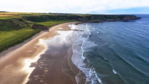 An aerial view of a coastal landscape with a sandy beach, grassy areas, and cliffs.