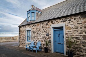 Un bâtiment en pierre avec une porte bleue et des volets bleus, un petit porche avec des chaises bleues et une enseigne indiquant « Sea Crest 219 ».
