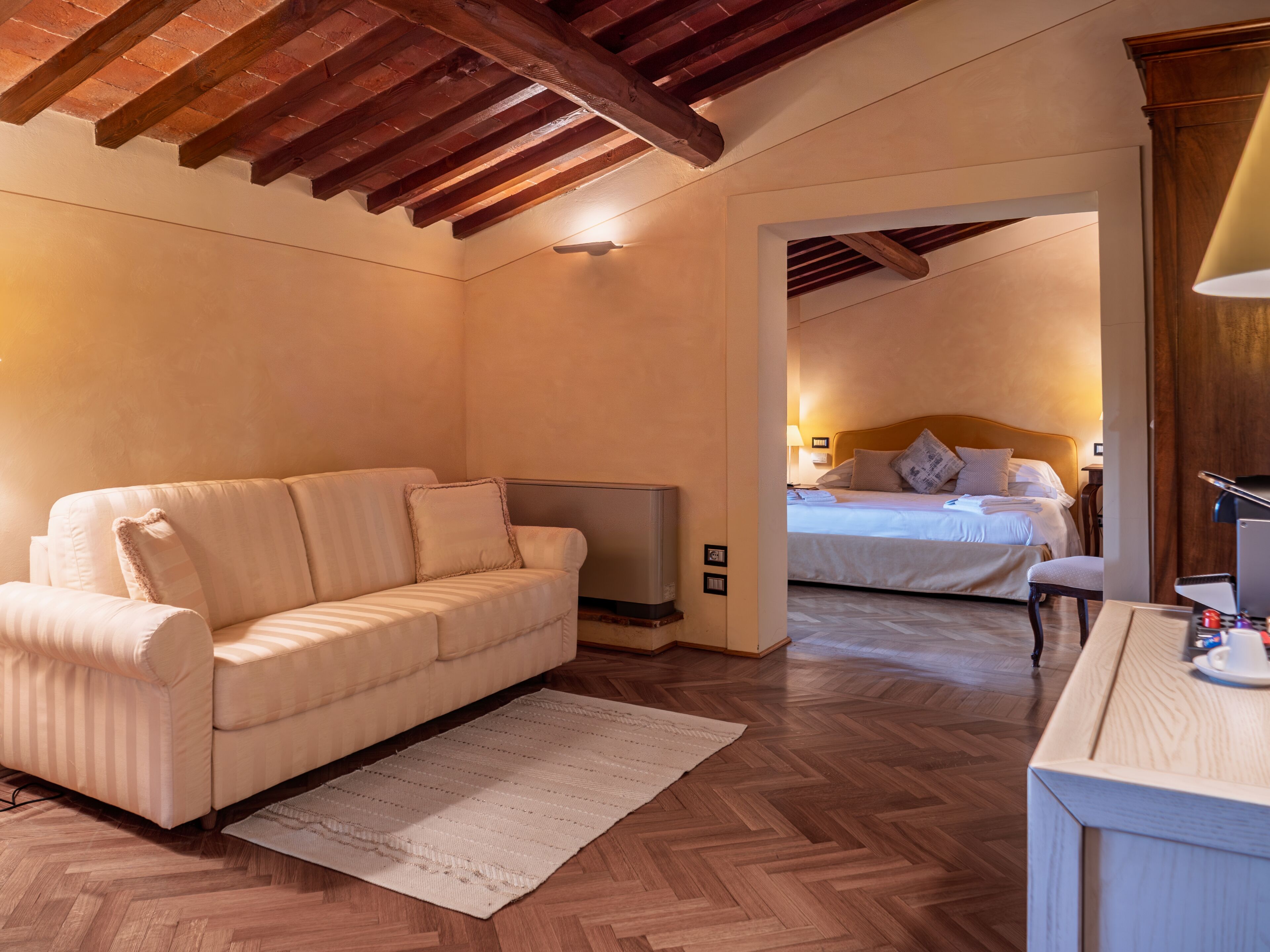 A hotel room with a sofa, a TV, a bed with pillows, a desk with a lamp, and wooden flooring.