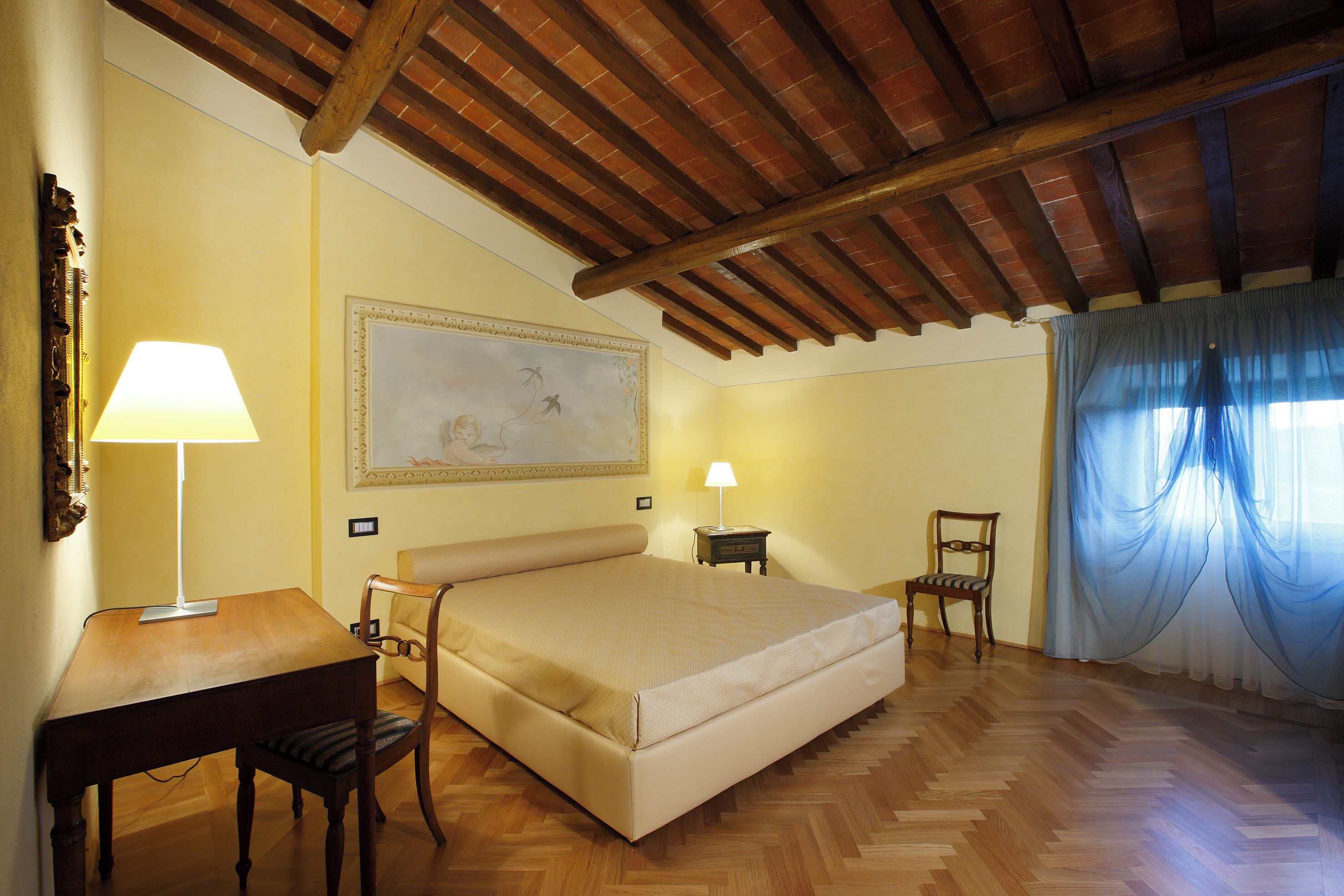 Quarto com teto de madeira, cama, escrivaninha, cadeira, mesa de cabeceira e uma pintura grande na parede.