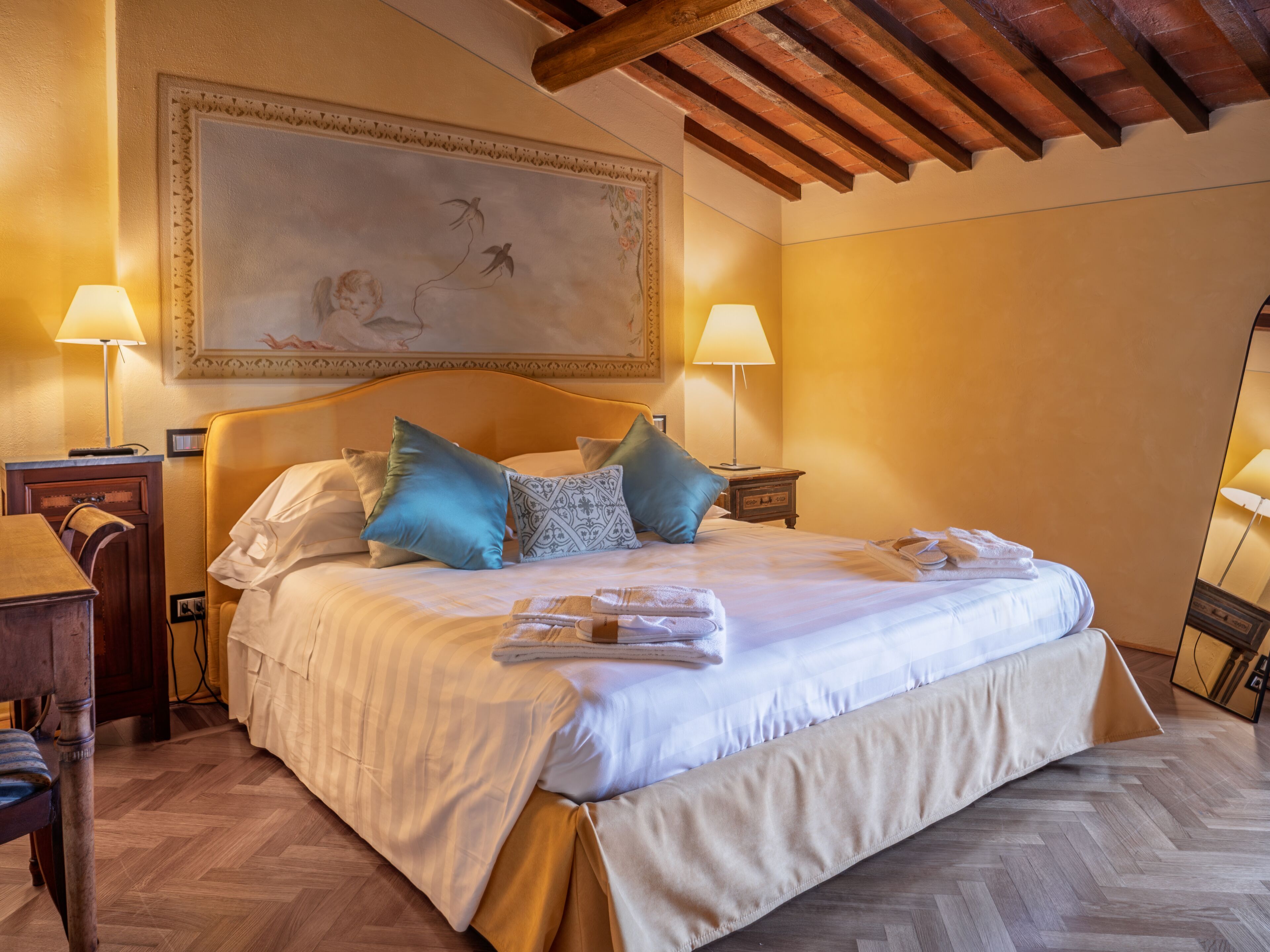 Une chambre d’hôtel avec un grand lit, deux tables de chevet, une chaise en bois, un bureau et un grand tableau accroché au mur.