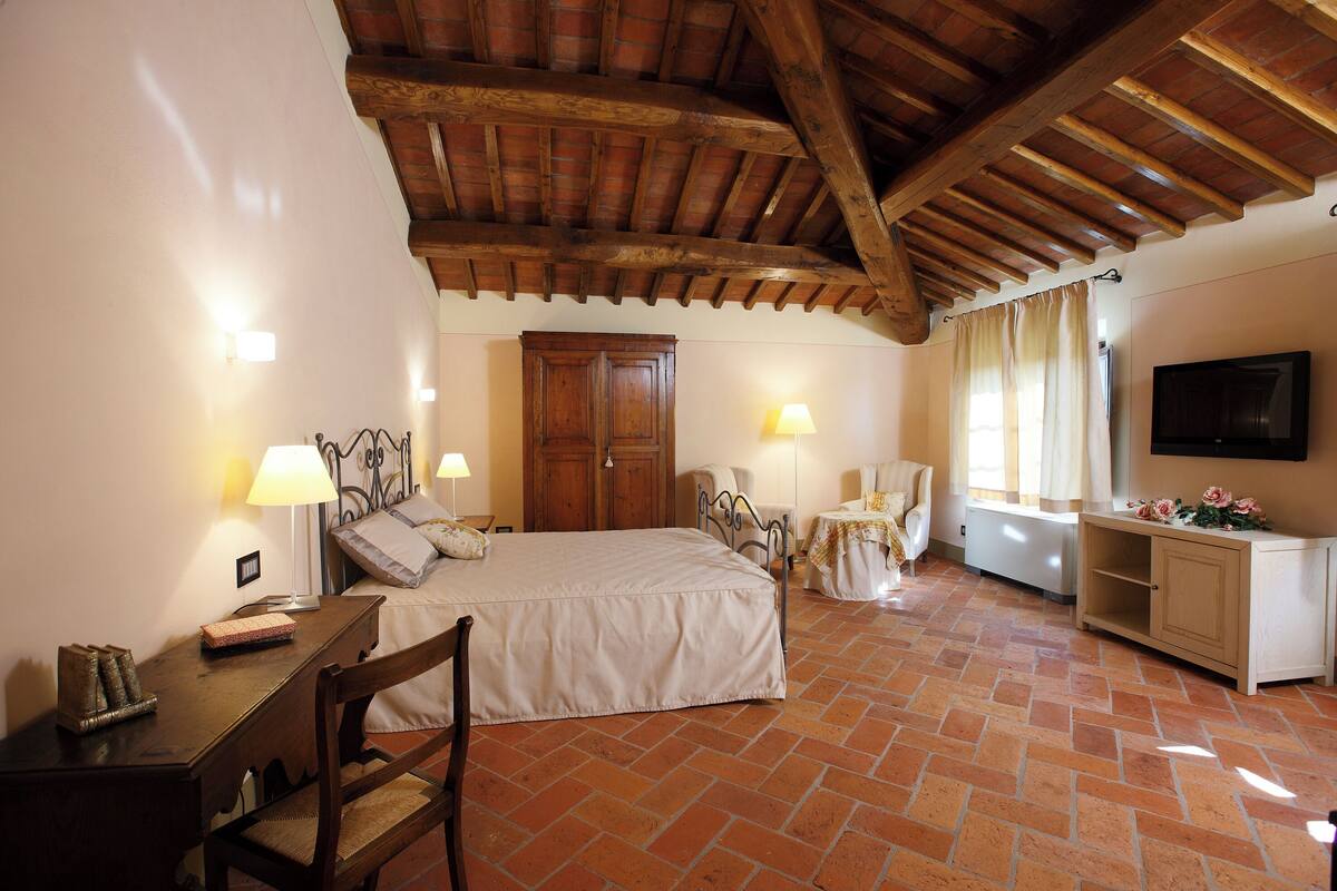 Un dormitorio con cama, mesitas de noche, escritorio, silla, armario, televisor y ventana con cortinas.
